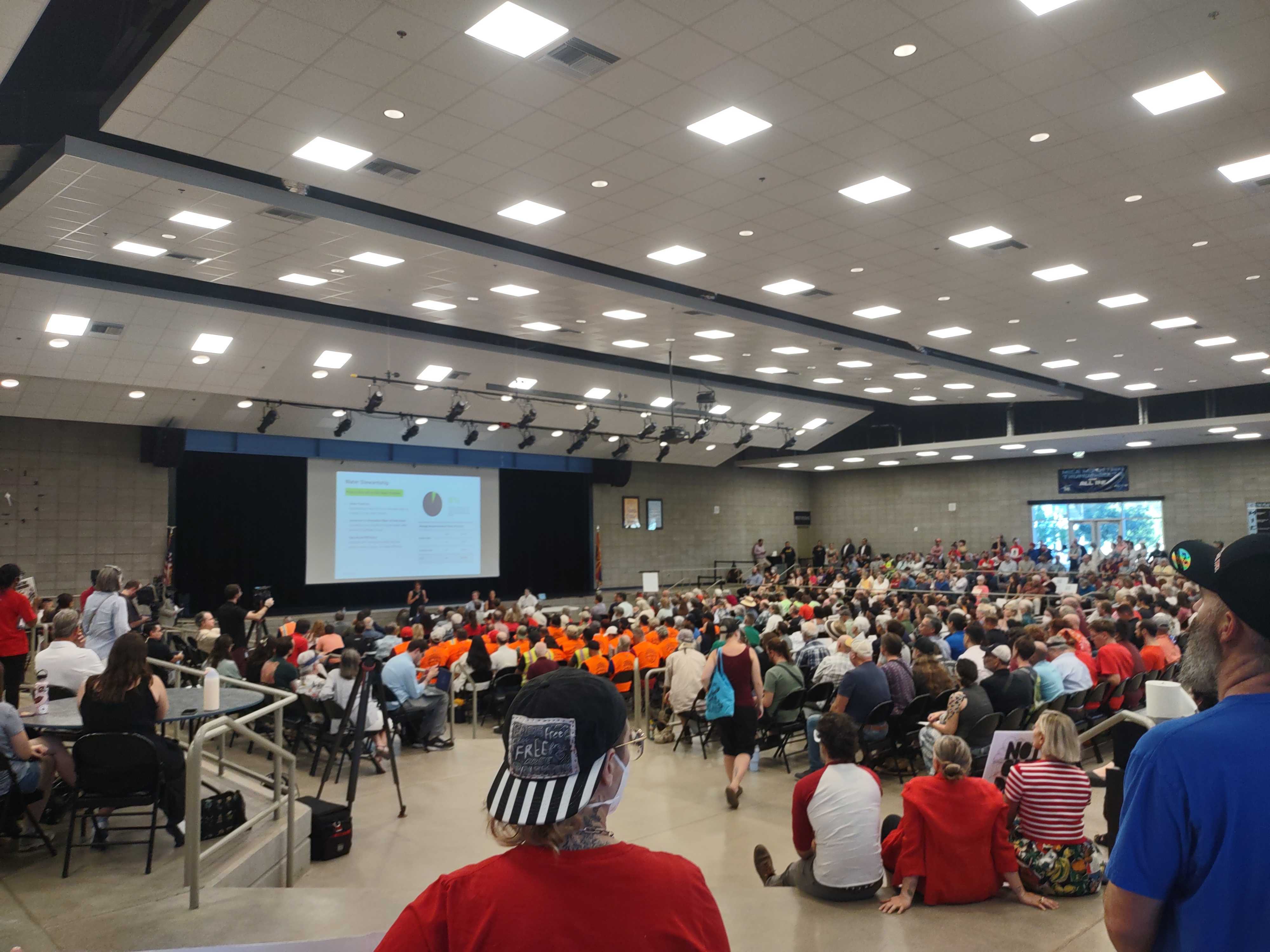 Scene from a city-wide Tucson meeting on Project Blue. Hundreds of participants pack chairs and sit on the floor facing a projector screen displaying data.