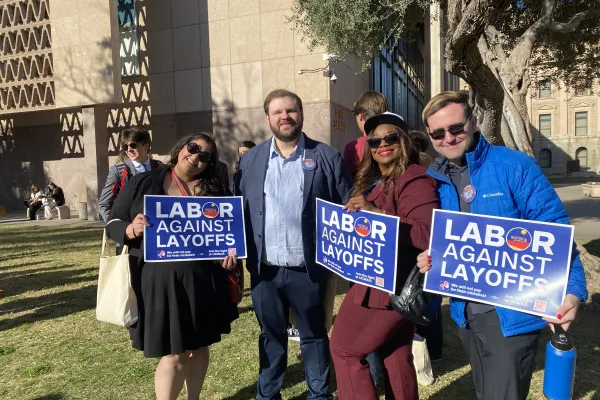 Picture of four people outside the Arizona State capitol. Three are holding blue signs that read "Labor Against Layoffs".