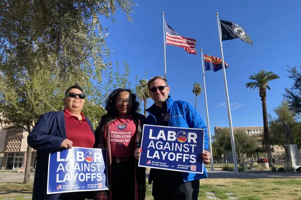 Three people in front of a US flag, an Arizona state flag, and a POW-MIA flag. Two of the people hold blue "Labor Against Layoffs" signs.