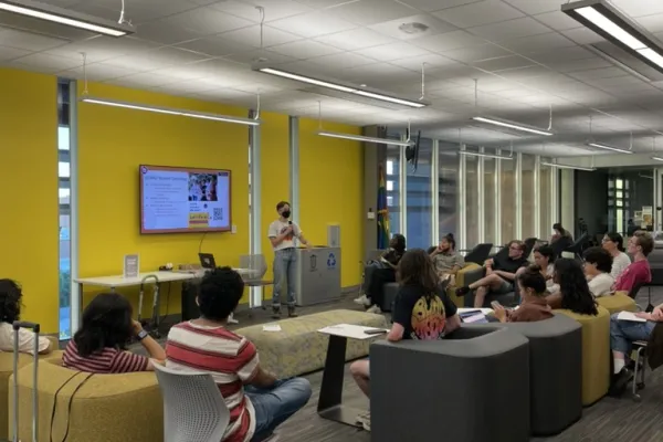 Person presenting to a large group of people indoors in a yellow room using a small TV on the wall. The crowd is mostly seated on puffy plastic chairs.