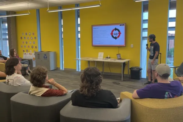 Person presenting indoors in a yellow room to a group of people seated on puffy plastic chairs.