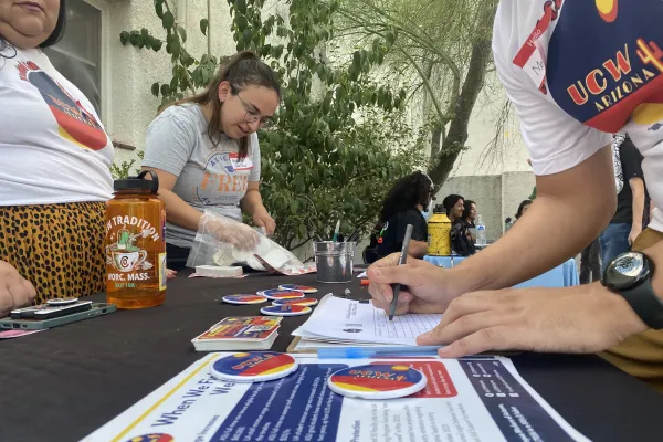 A person signs up for UCW in front of people wearing UCWAZ shirts. The shot features a zoomed-in view of a table with flyers, pins, buttons, stickers.