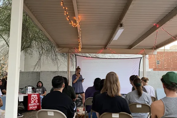 Person presenting outdoors on a projector screen under a roof decorated with twinkle lights. The person is holding a microphone and speaking to a group of people facing away from the camera, seated on gray metal folding chairs.
