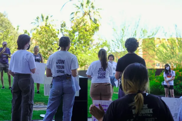 The backs of several people standing on stage during a rally/press conference. Two of them wear white tshirts that read "Join Your Union" in blue.