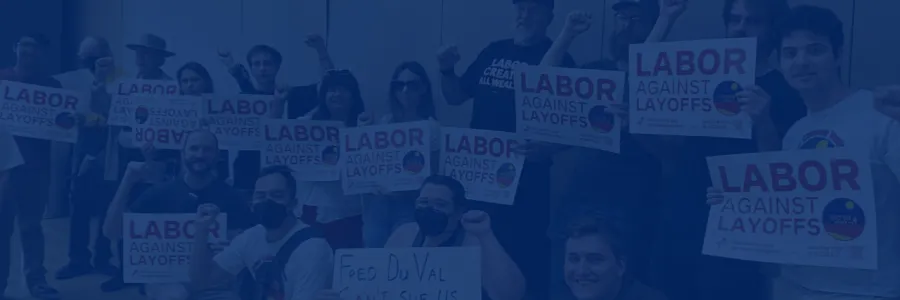 People standing in a group indoors, all smiling, holding up fists, and holding up signs that say "labor over layoffs"