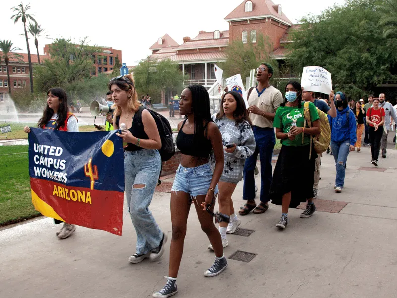 Group of people marching in a line on campus. Front two people are holding a UCWAZ-branded banner