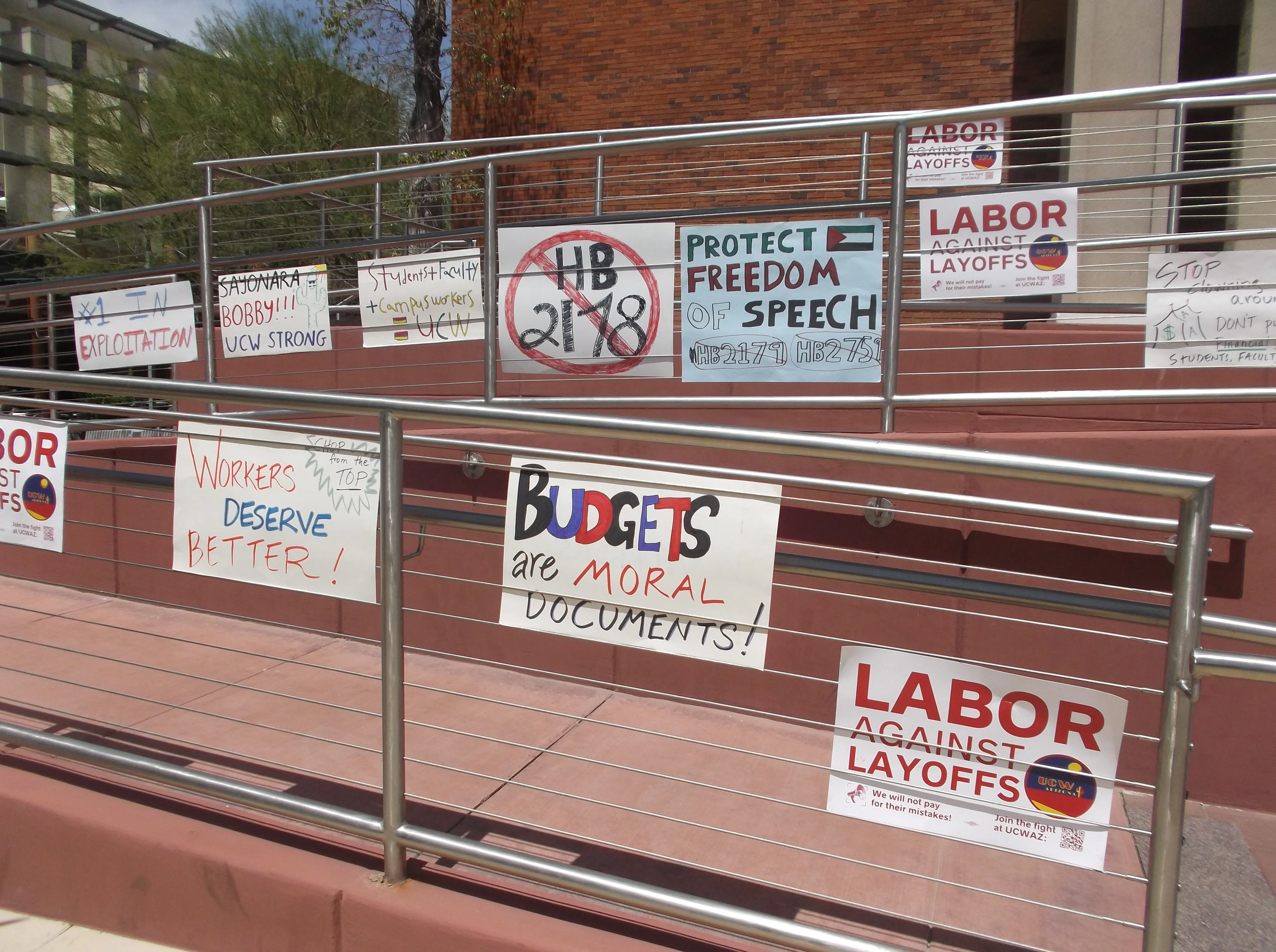 A series of signs on a railway with a fence bordering it. Signs include "protect freedom of speech," "labor oagainst layoffs," "no to HB 2178"