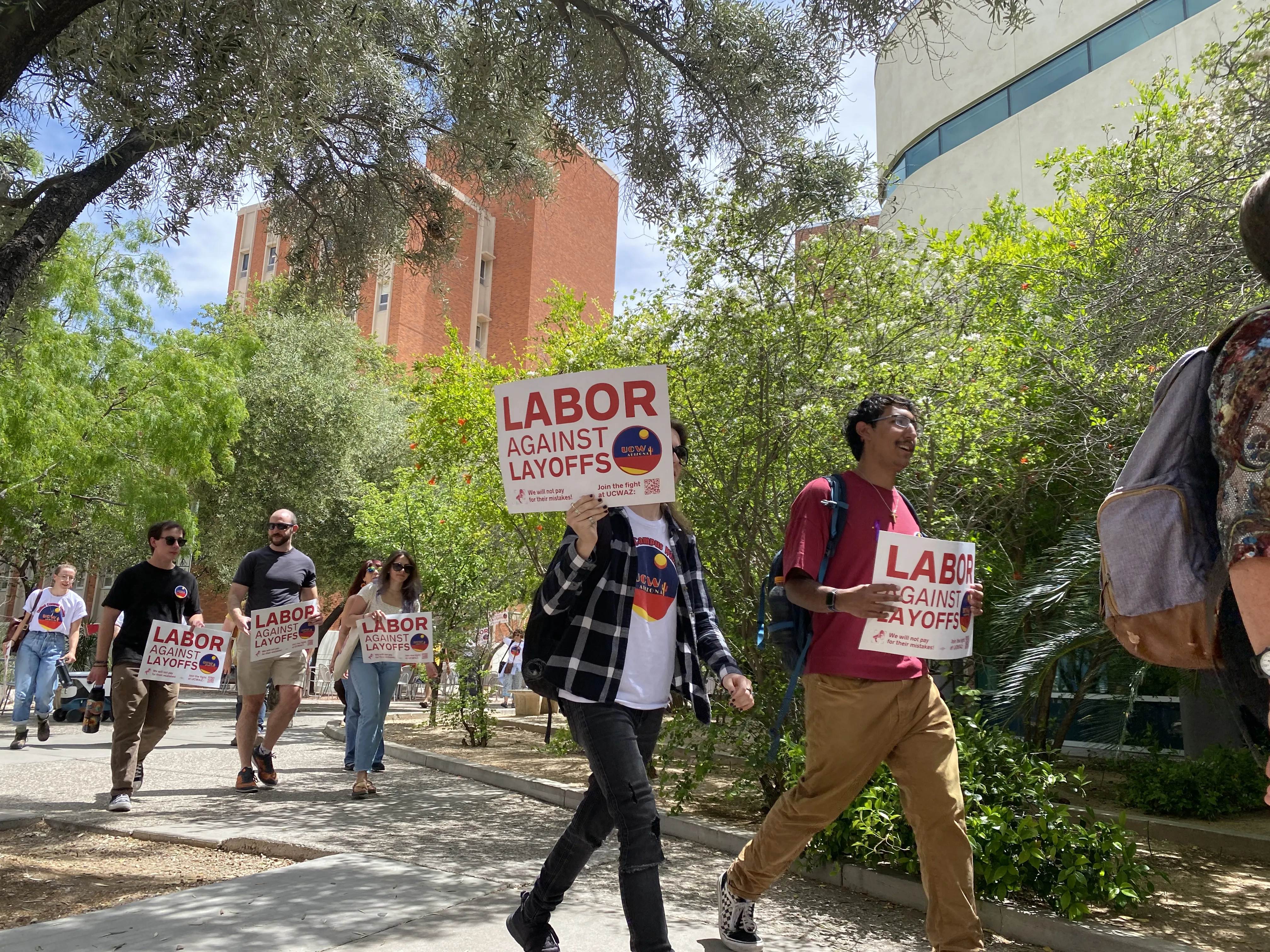 People walking outside at UA. Background is luscious green trees. People are holding "labor against layoffs" branded UCWAZ signs.