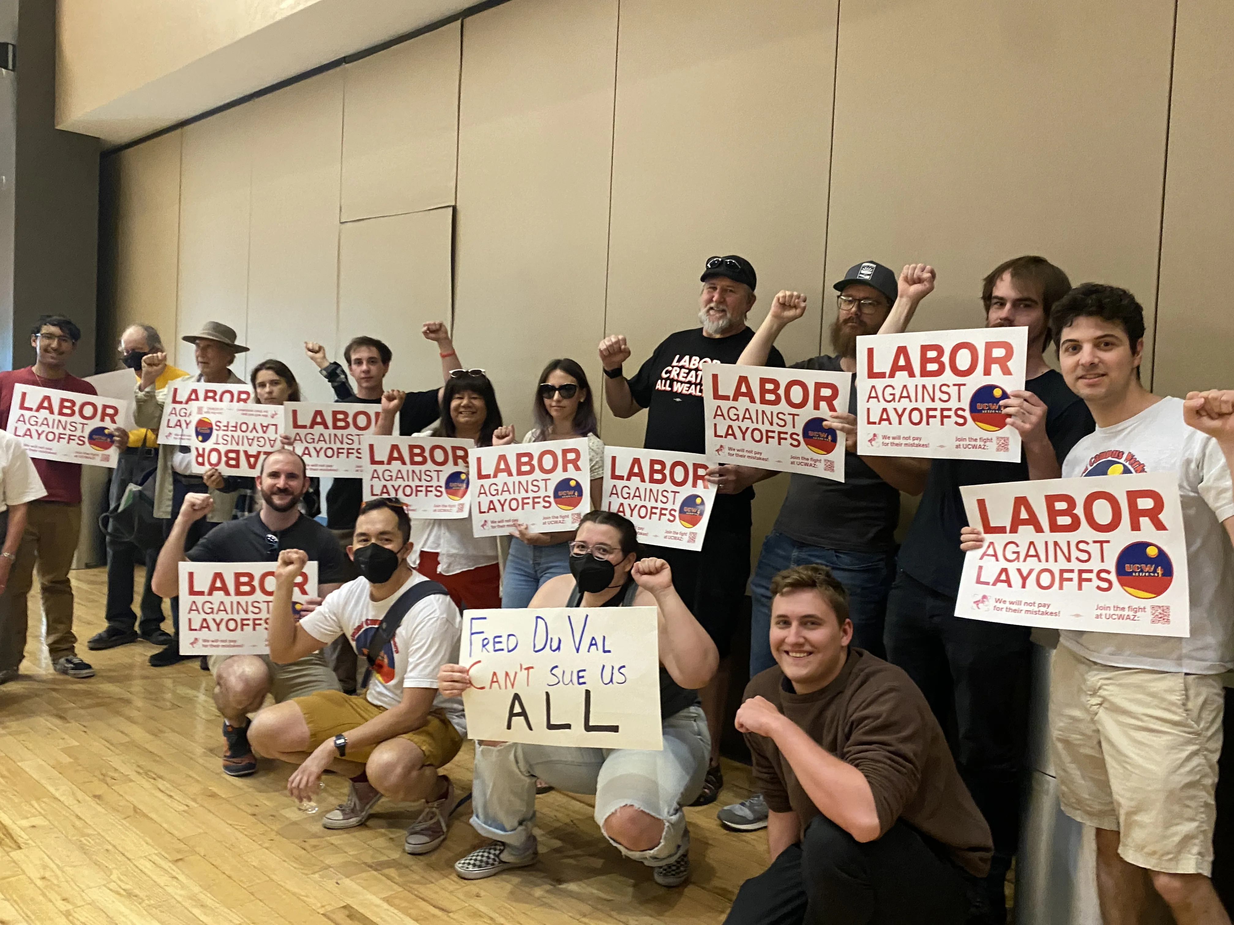 A group of people sitting in a room with their fists up, holding signs that say "Labor against layoffs" on UCWAZ branded paper.