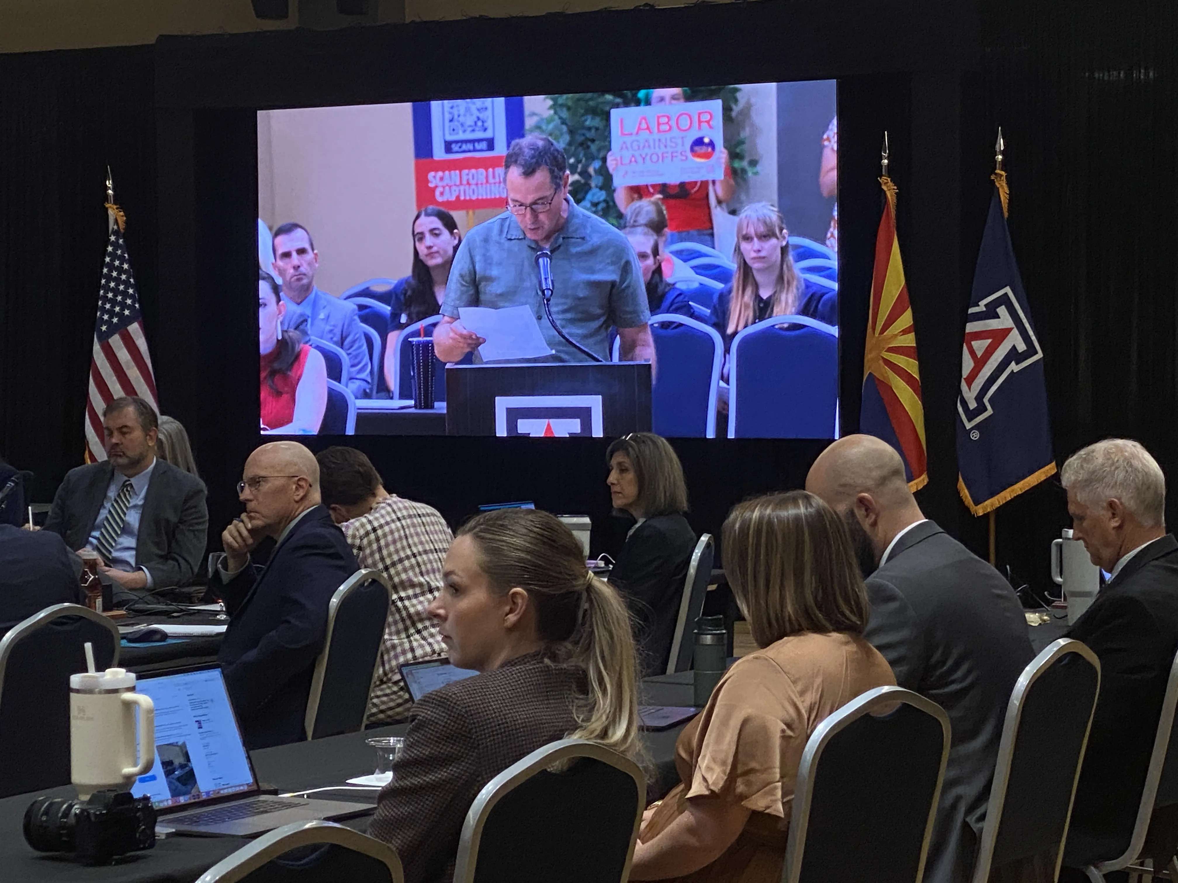 Image on a projector screen of a person speaking at a podium; in the foreground is a group of professionally-dressed individuals watching the screen and listening to the speaker. Blurry in the background is a "labor against layoffs" UCWAZ-branded sign.