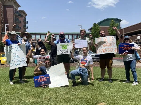 A row of people standing outside at NAU, holding signs reading "labor against layoffs" and "let the students speak" and "end student repression", all holding up solidarity fists.