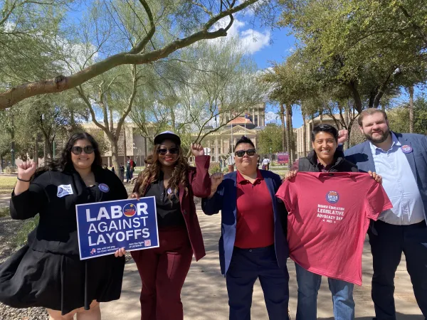 Picture of four people outside the Arizona State capitol. Three are holding blue signs that read "Labor Against Layoffs".
