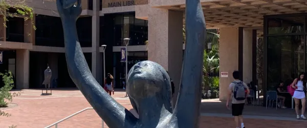 Bronze statue of a woman raising her arms up to grab some birds just out of the shot. The statue is pictured in front of the University of Arizona's main library in the sun.