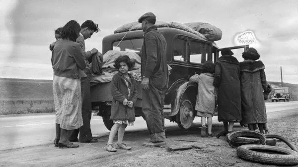 A black and white image of a Mexican family loading a car on a highway shoulder in California. A child in the foreground looks at the camera concernedly.