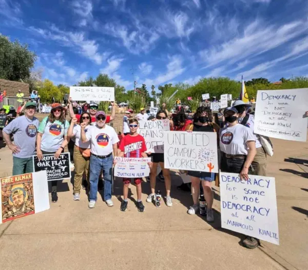 A group of people standing outside in the sun, wearing union tshirts and holding signs with May Day slogans on them.