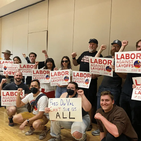 Group of people standing in a group, holding up solidarity fists and "labor over layoffs" signs