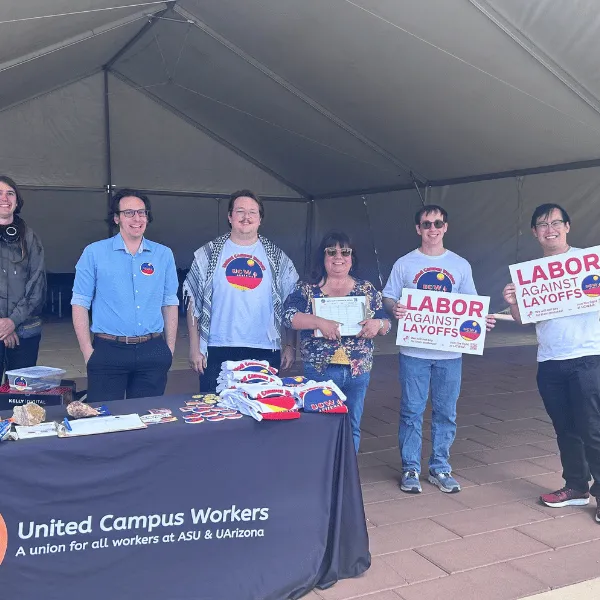 Group of people standing behind UCWAZ branded table, holding "labor against layoffs" signs