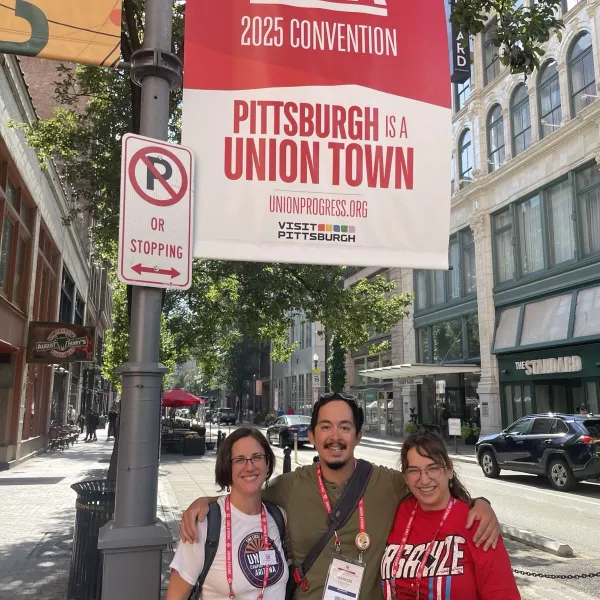 Three people underneath a "Welcome CWA 2025 Convention" flag. One wears a UCWAZ tshirt.