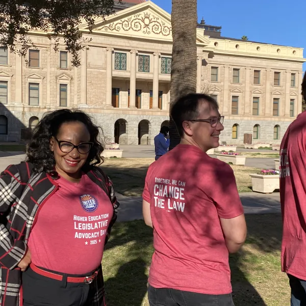 Three people stand outside the capitol building in Phoenix, AZ. They all wear red tshirts. The front reads "Higher Education Legislative Advocacy Day 2025". The back reads "Together we can change the law".